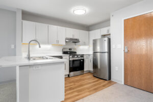 Bright modern kitchen with white cabinets, stainless steel fridge and oven, under-cabinet lighting, and a wooden door on the right.