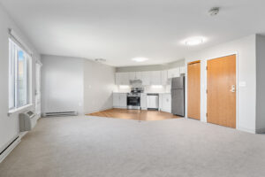 Open-concept kitchen and living area with white cabinets, stainless steel appliances, wood doors, and large left-side windows.