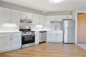 Bright modern kitchen with white cabinets, stainless steel appliances (fridge, oven, dishwasher), and light wood flooring.