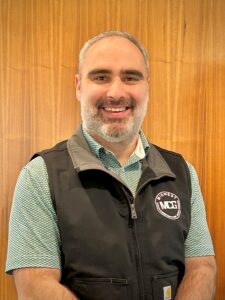 Smiling man with short gray hair and beard, wearing a black vest over a green patterned shirt, standing against a wooden wall with a Midwest logo patch on the vest.
